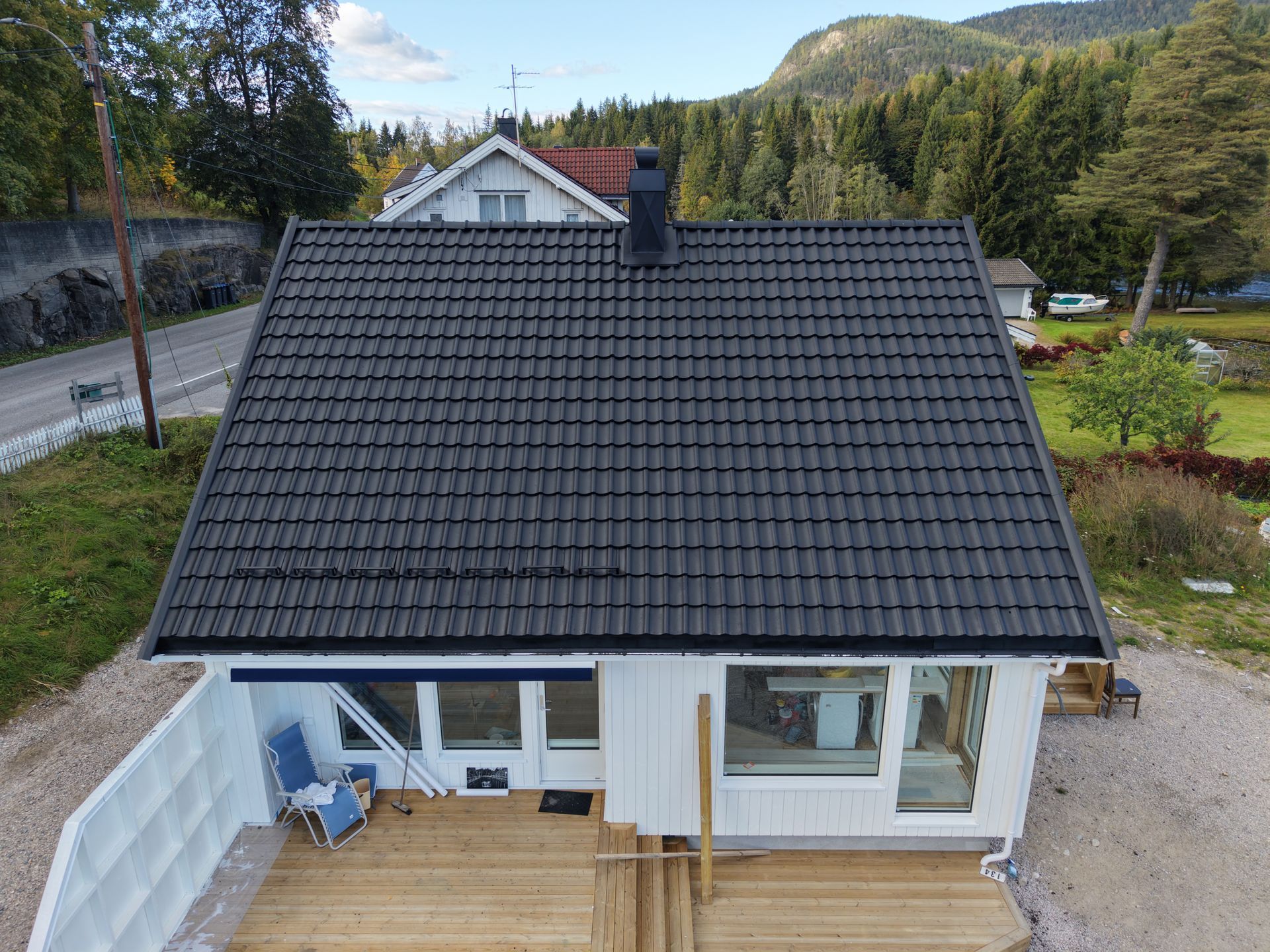 Luftfoto av et hvitt hus med svart tegltak og en treterrasse, mot et bakteppe av trær og en ås.