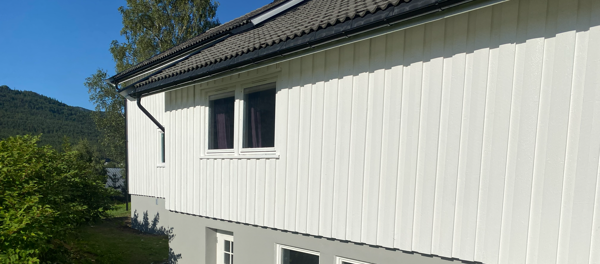 Sidevisning av et hvitt hus med vertikale sider, grått fundament, mørkt tegltak og to vinduer mot en blå himmel.