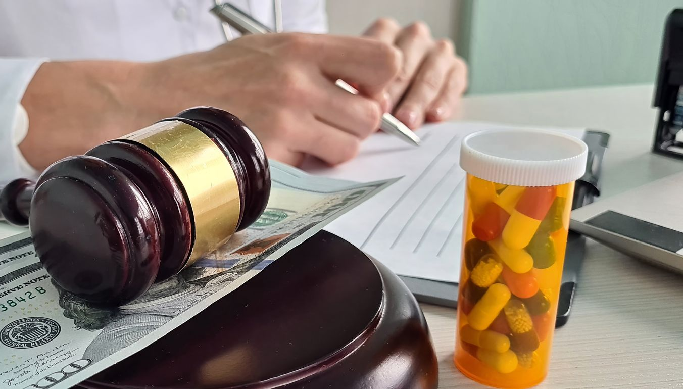 Judge’s gavel and case files beside a hand signing paperwork, with a pill bottle in the foreground.