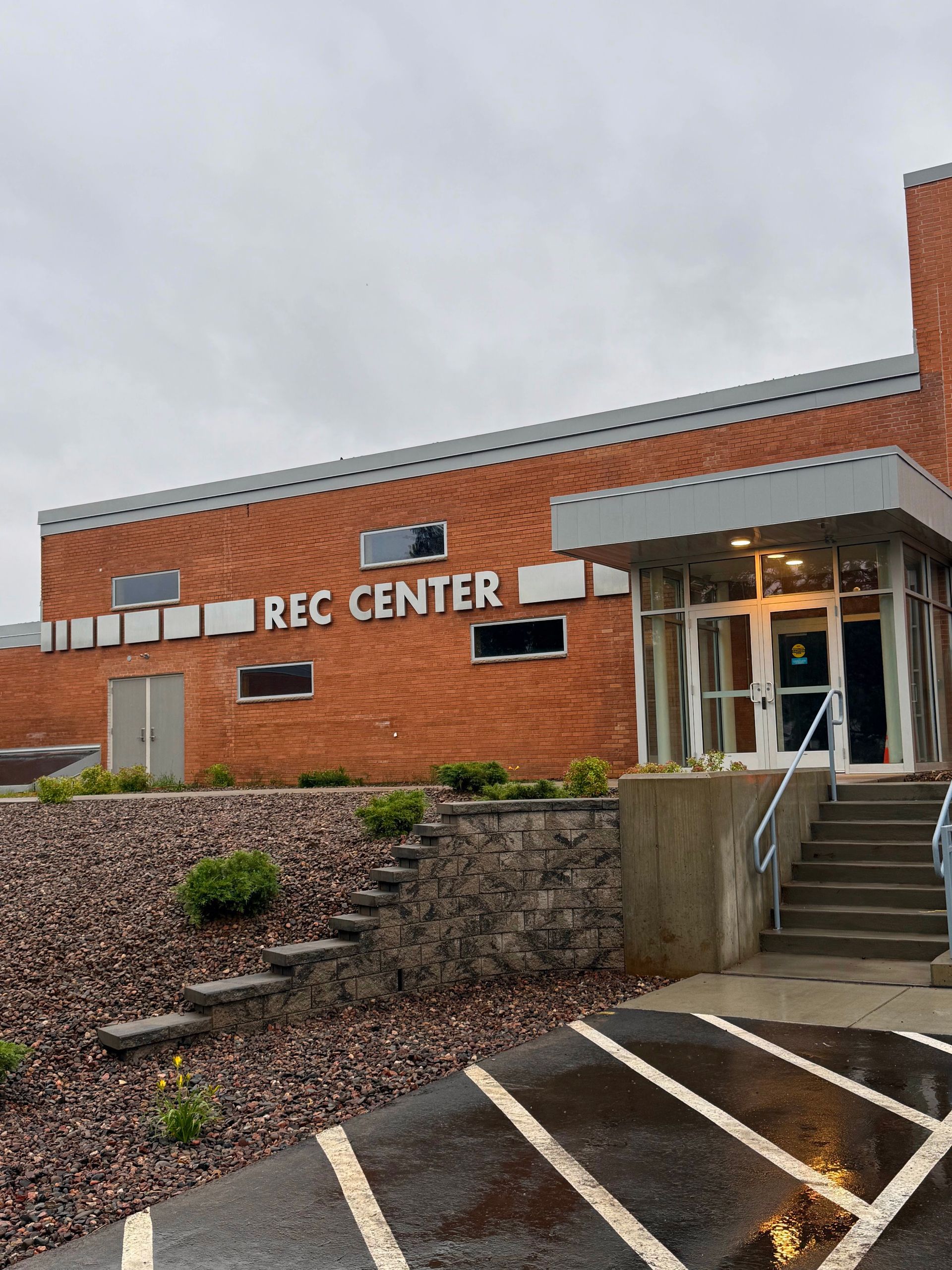 Brick recreation center entrance with stairs, ramp, and reserved parking spaces on a cloudy day