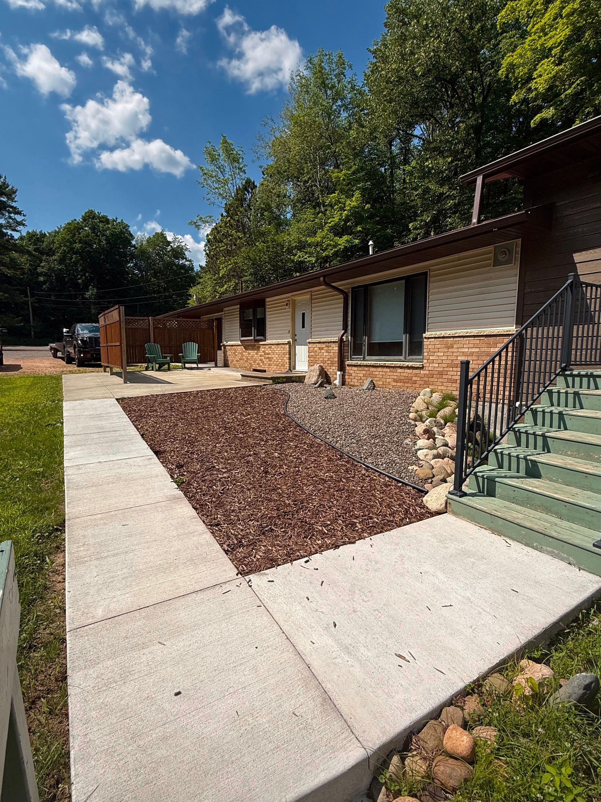 Concrete sidewalk by a brick building with green steps, landscaping rocks, and trees under a blue sky