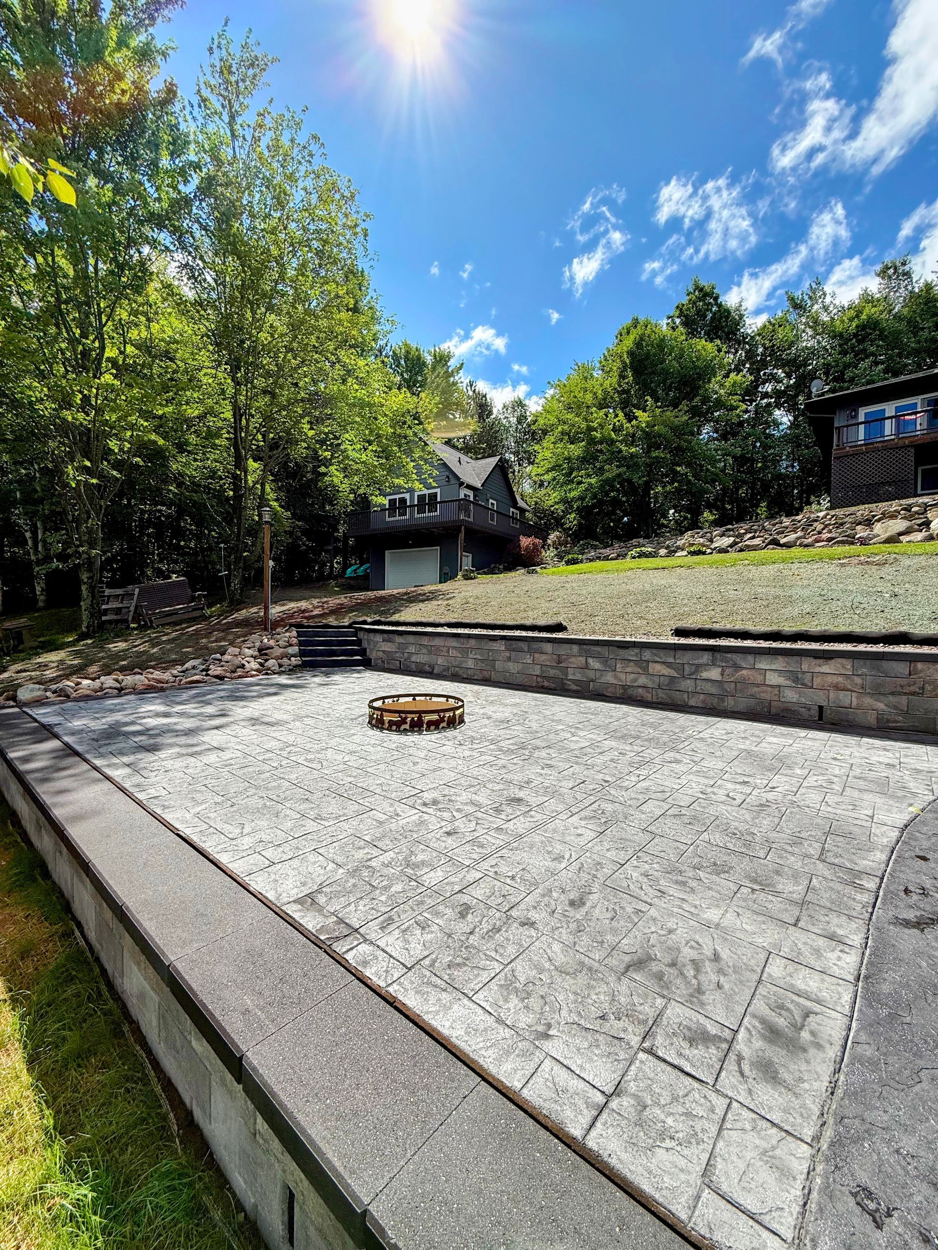 Sunny backyard with a gravel patio, stone retaining wall, and steps leading to a tree-lined yard.