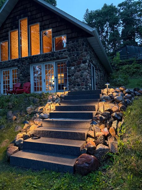 Stone cottage at dusk with warm-lit windows, front steps, and garden path surrounded by flowers and rocks