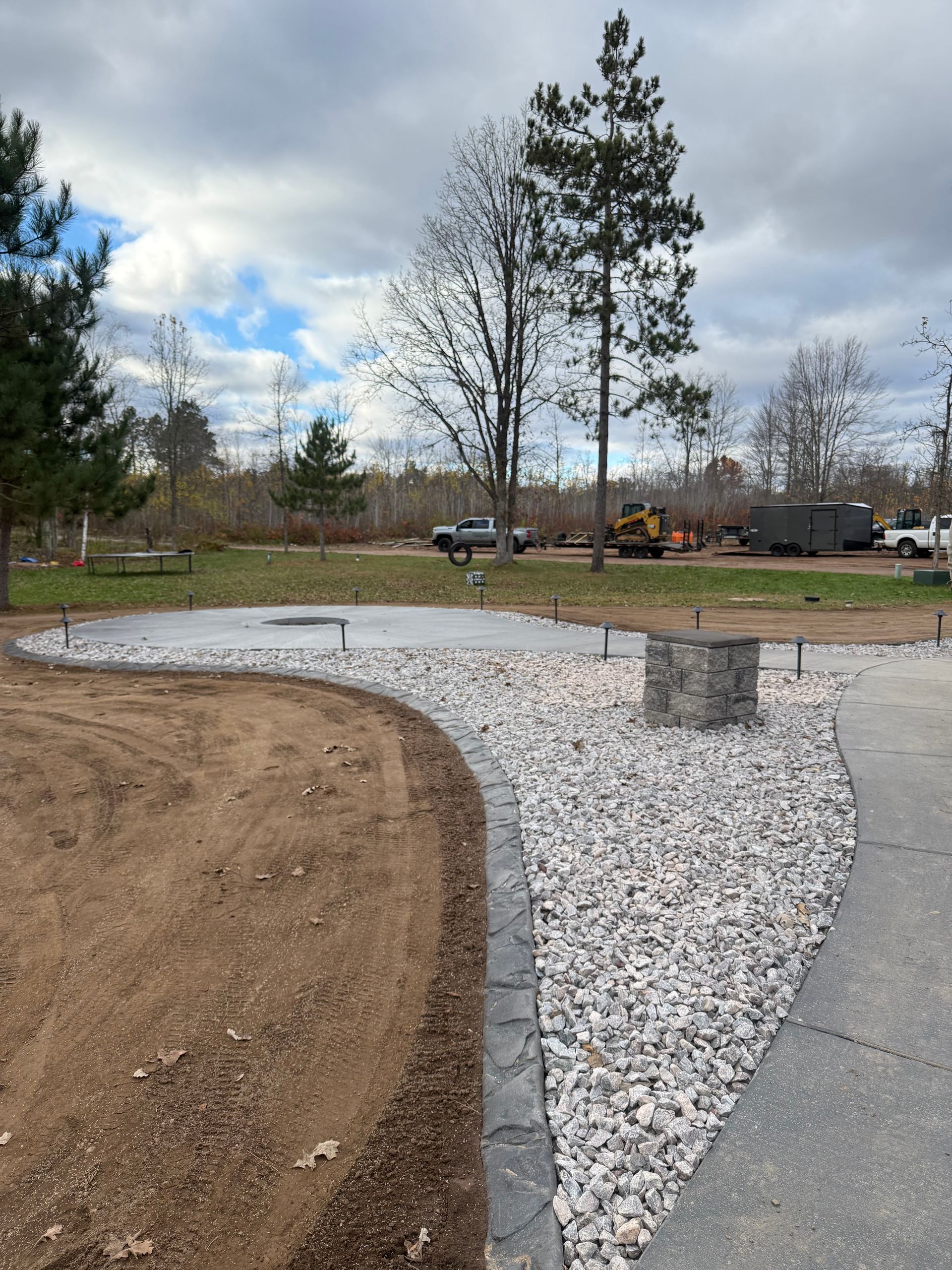 Landscaped yard with gravel path, curved border, stone bench, and trees under cloudy sky.