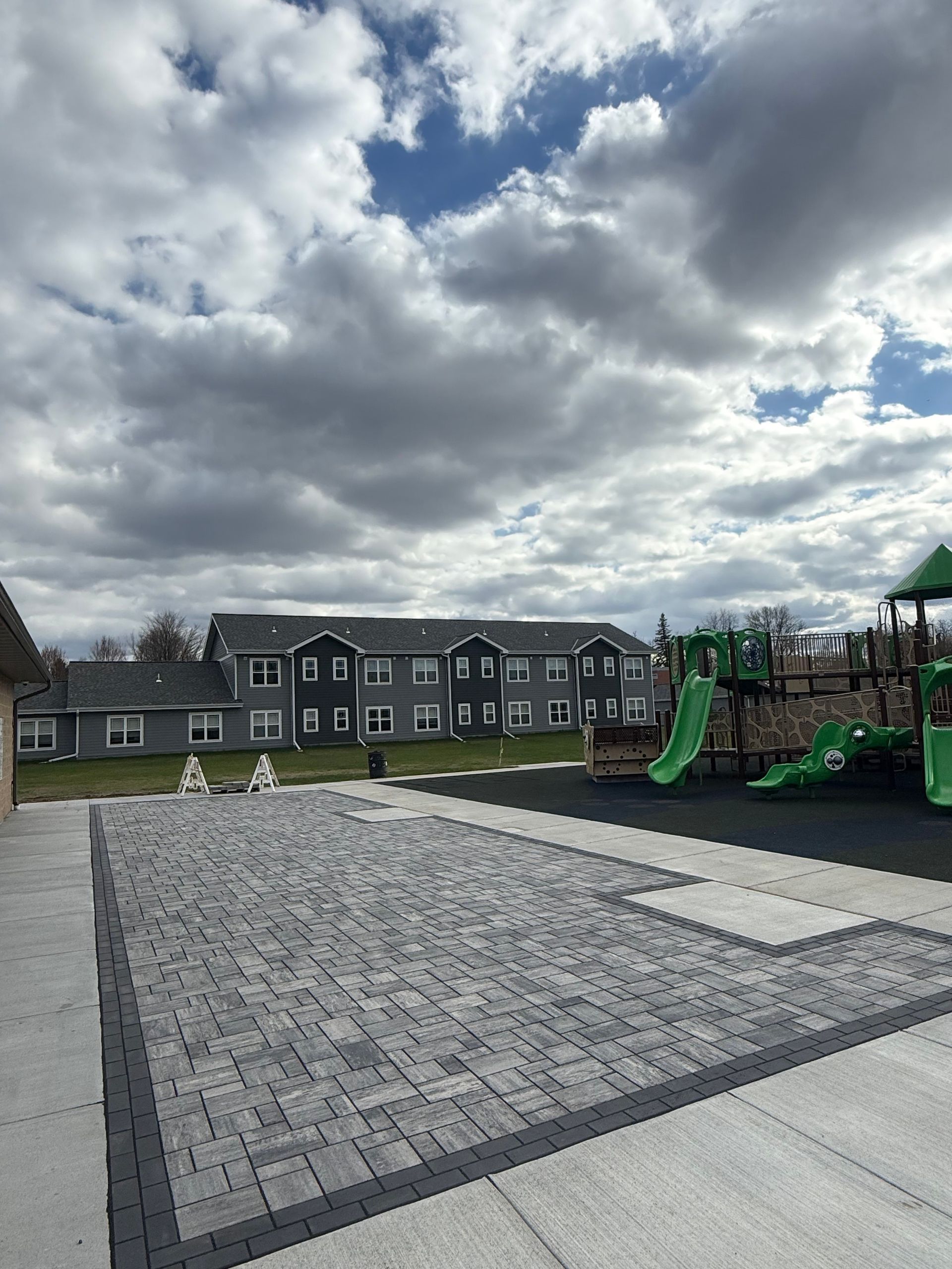Empty playground with green play structure, paved area, apartment buildings, and cloudy sky.