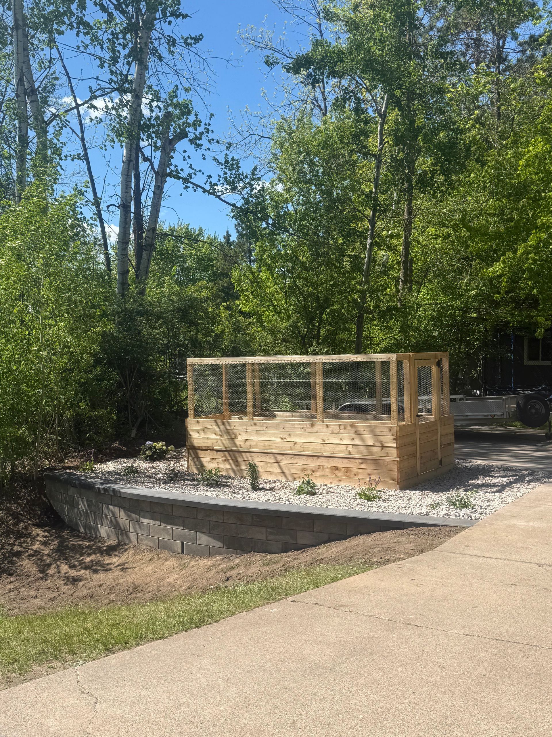 Wooden raised garden bed beside a concrete path, surrounded by trees and rocks.