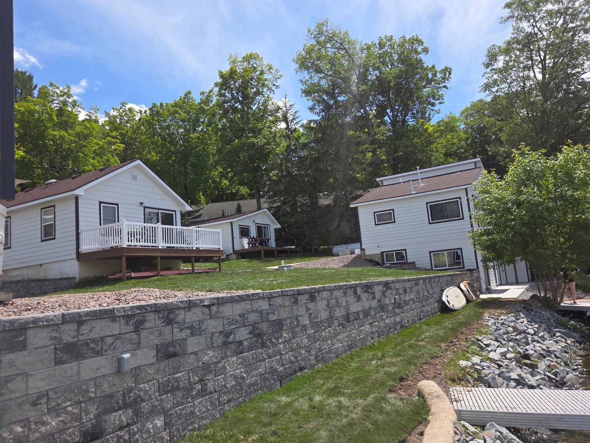 Stone retaining wall by a gravel path below two white houses on a wooded hillside under a blue sky