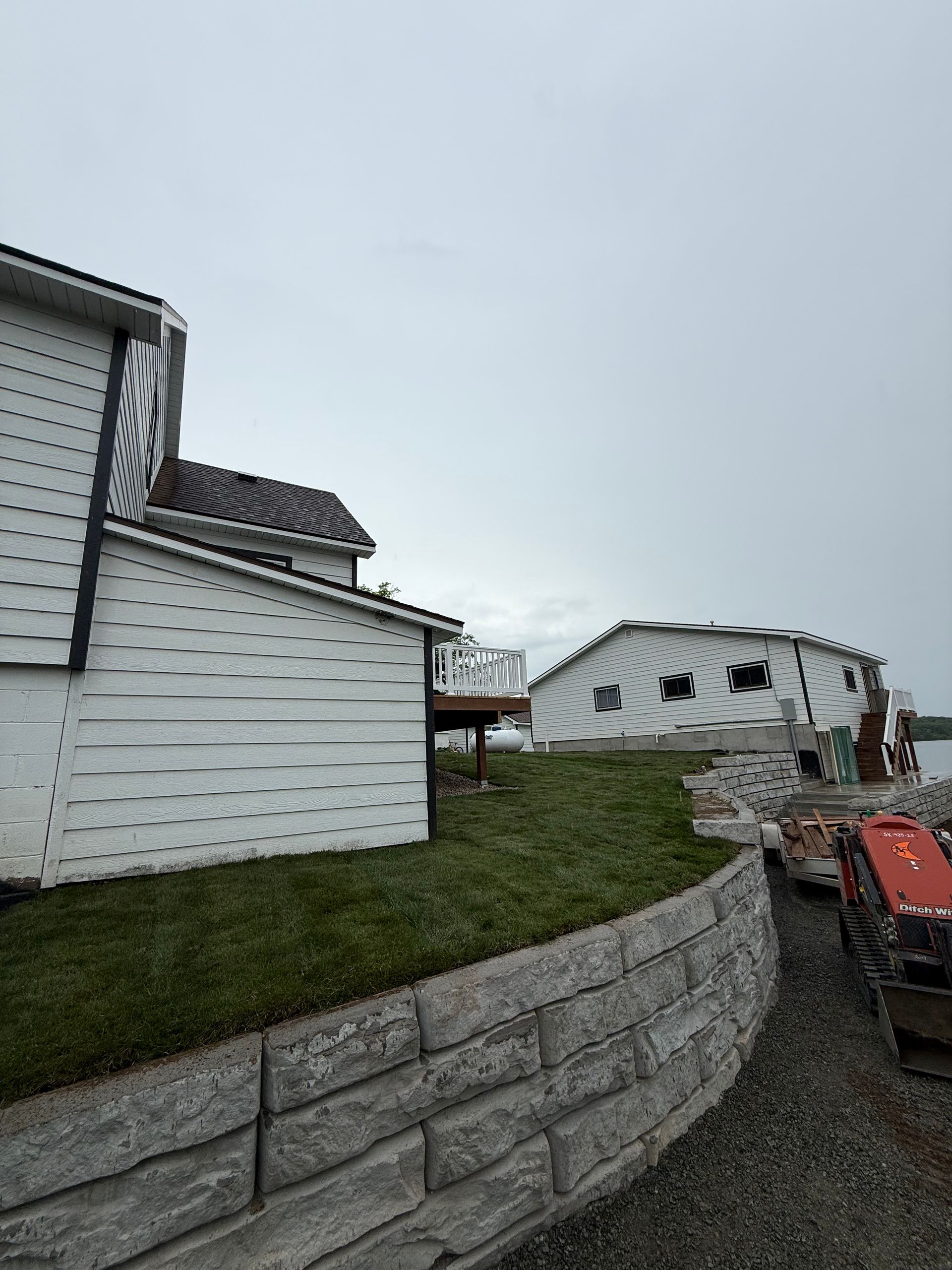 White house with siding, retaining wall, and damaged roof near an overturned white trailer in a gravel yard