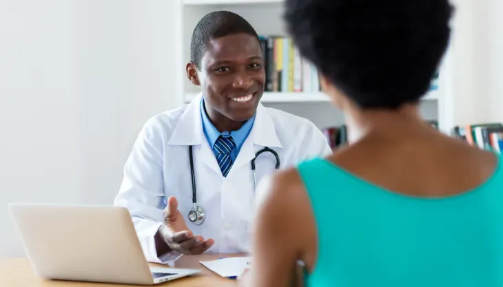 A doctor is talking to a patient while sitting at a desk with a laptop.