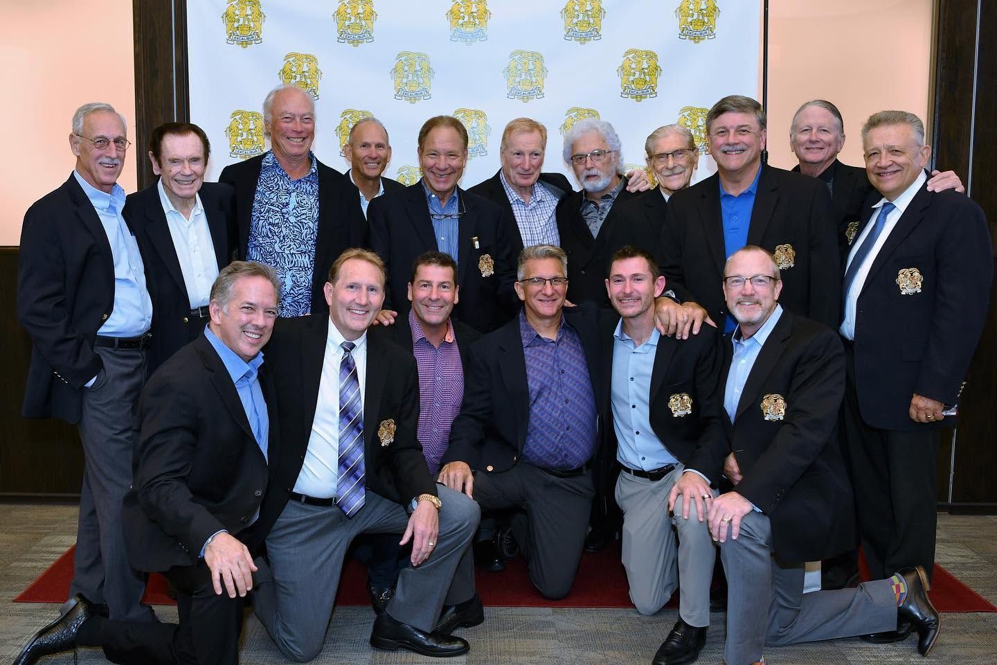 Group of men posing in suits at an indoor event, smiling in front of a patterned backdrop.