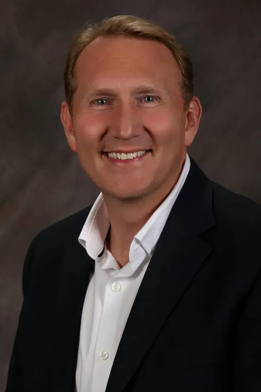 Studio headshot of a smiling man in a black blazer and white shirt on a dark background