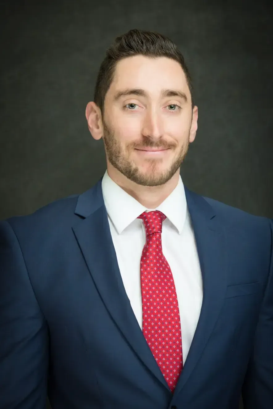 Studio headshot of a man in a navy suit, white shirt, and red tie against a dark background