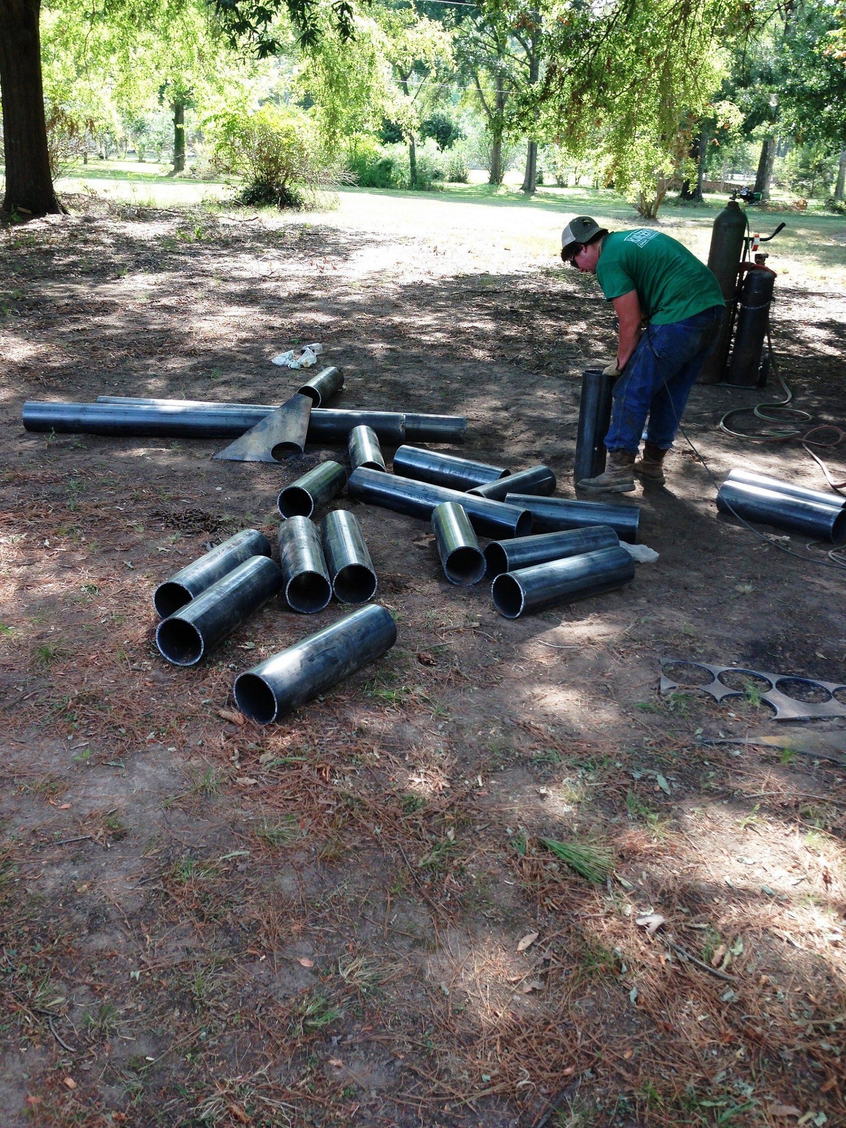 Man in green shirt arranging black pipes on a grassy, wooded area.