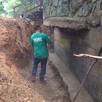 Man in green shirt measures a wall in a trench beside a stone building.