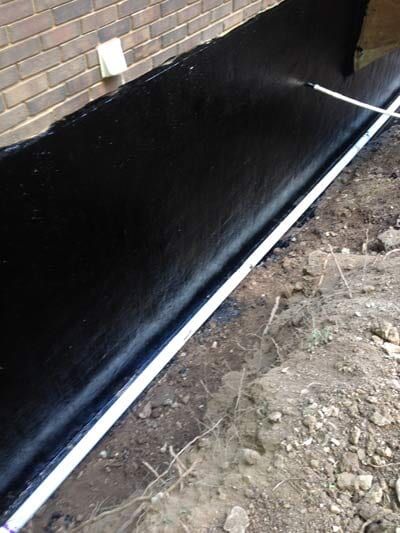 Black waterproofing coating being sprayed onto a foundation next to a brick wall.