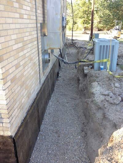 A trench beside a brick building, filled with gravel, exposing the foundation. Electrical boxes are mounted on the wall.