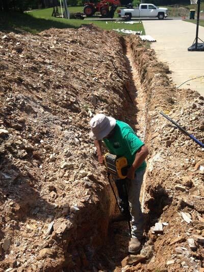 Man in green shirt uses a jackhammer to dig a trench near a driveway.