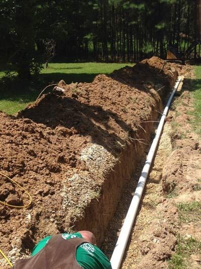 A trench with a white pipe being installed, with a person working in the foreground, outside.