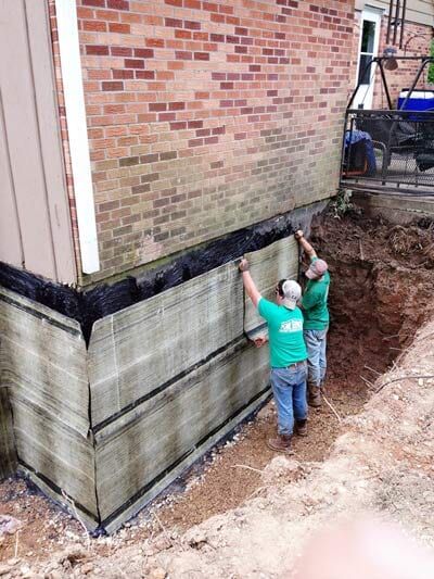 Two workers install foundation waterproofing on a brick building.