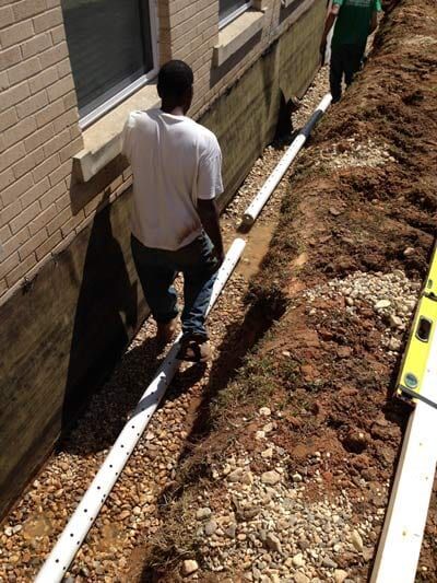 Workers installing drainage pipe along a building's foundation in a dirt trench.
