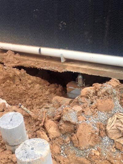 Hole in ground reveals plumbing under a structure; white pipe runs along the building.