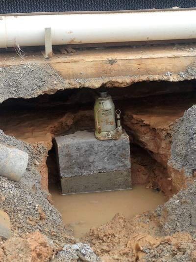 A hydraulic jack atop a concrete block in a water-filled excavation under a white pipe and damaged asphalt.
