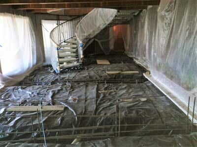 Interior room under construction, gray floor covered in plastic, spiral staircase, white walls.