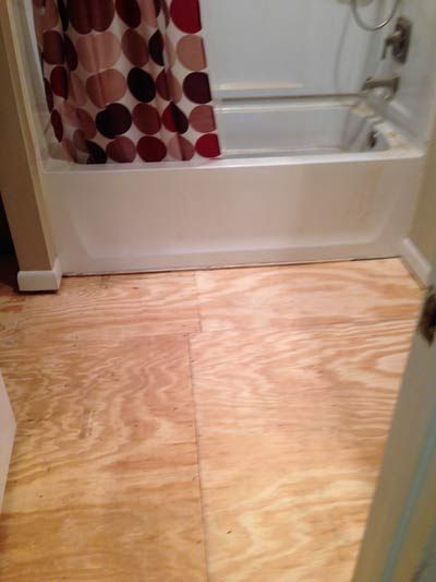 Plywood bathroom floor in front of a white bathtub with a patterned shower curtain.