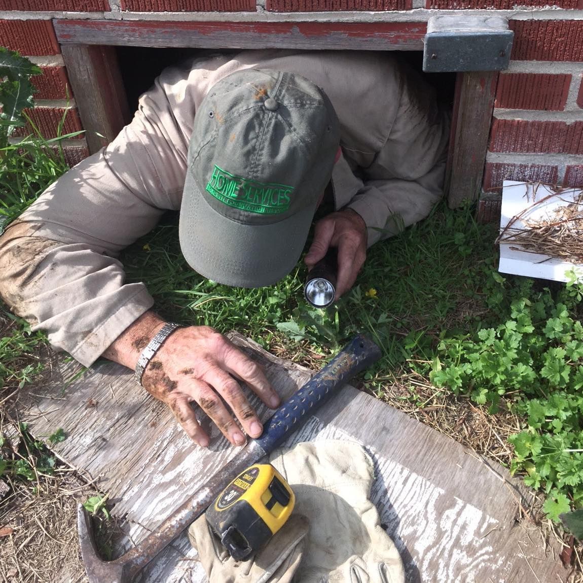Man crawls into small brick opening, holding a flashlight and tools. Green grass surrounds the entrance.