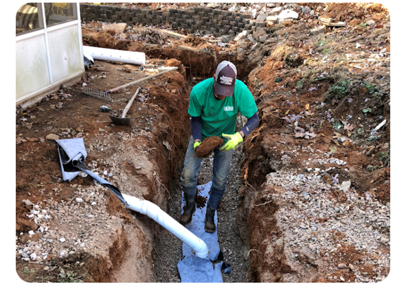 A trench dug in a yard with retaining walls, a backhoe in the background, and leaves scattered throughout.