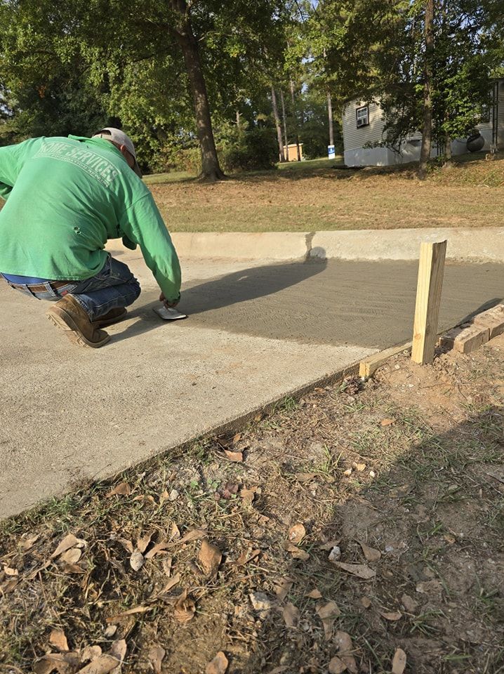 A person in green shirt smoothing concrete on a pathway with a tool outdoors, daytime.