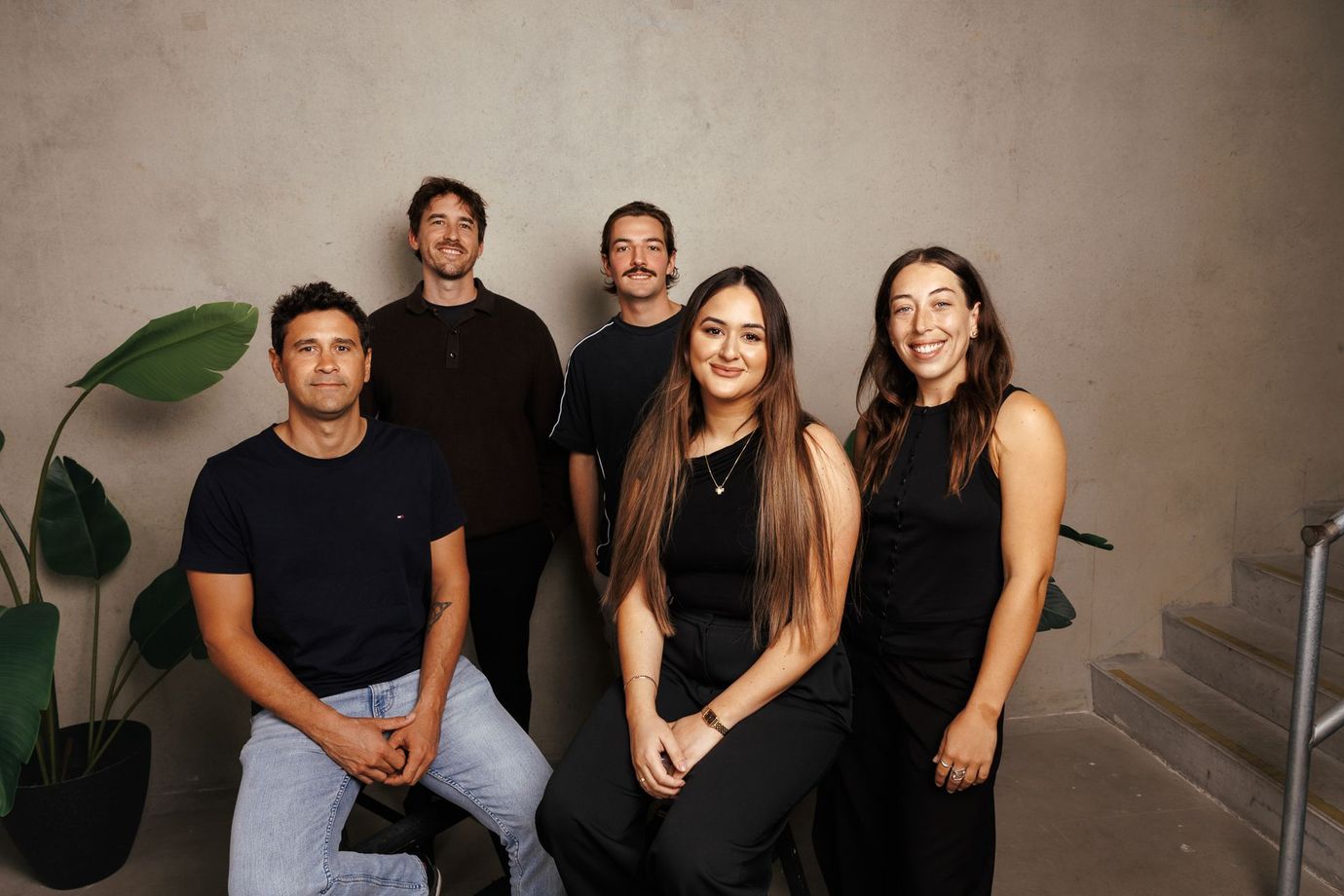 Five people posing together indoors on stairs, with a potted plant and beige wall behind them.
