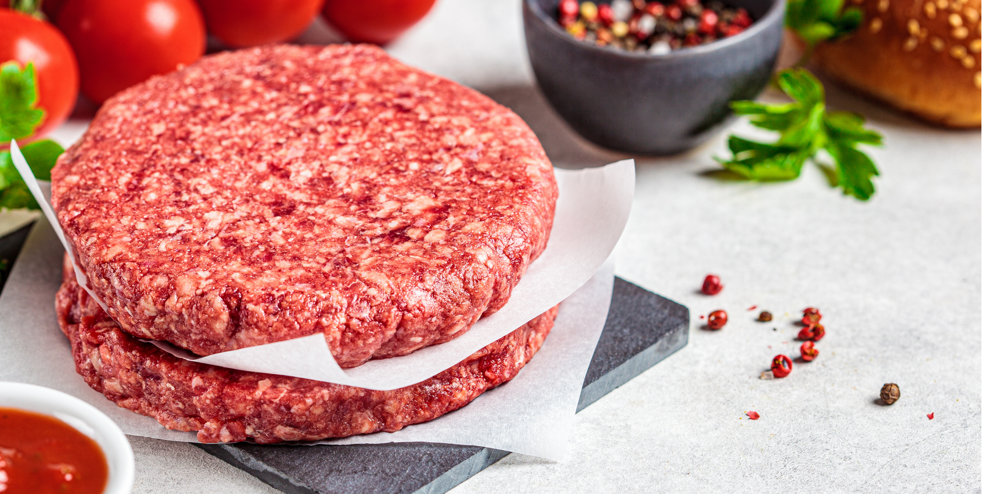 A raw, seasoned beef steak rests on a light-colored wooden cutting board scattered with peppercorns.