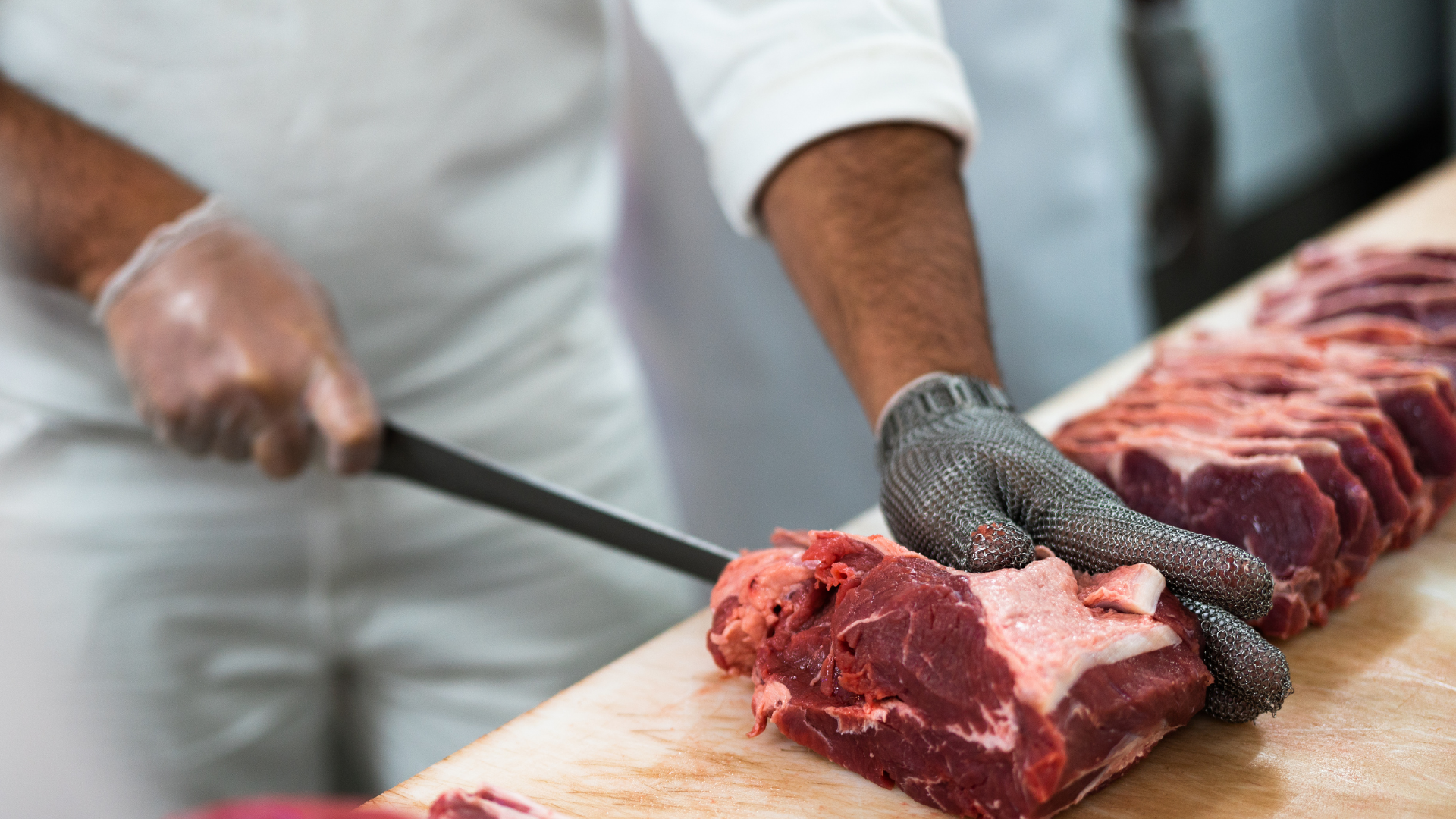 A butcher in a white coat wearing a protective mesh glove cuts a raw piece of beef on a wooden table.