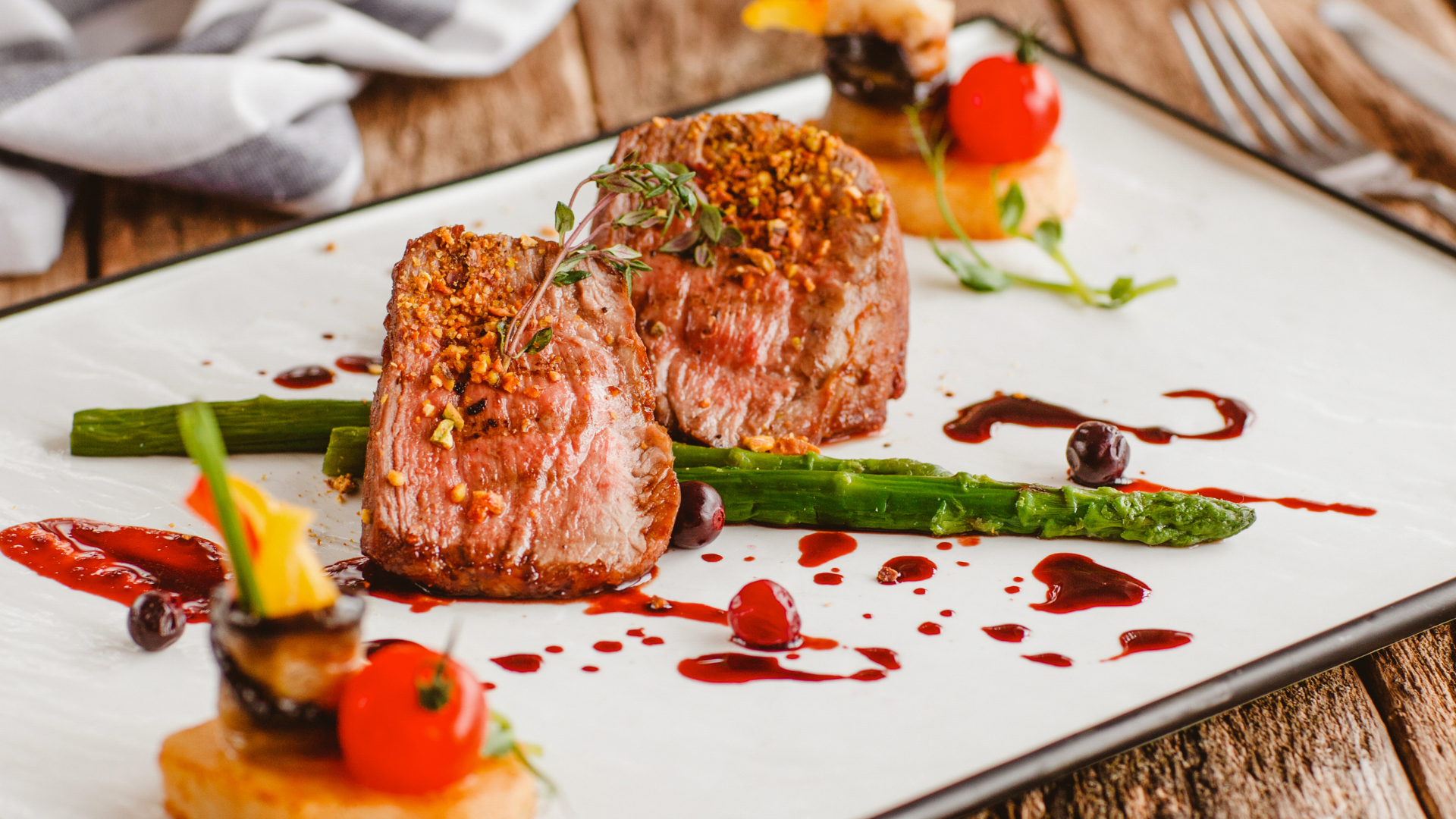 Two seared steak medallions topped with herbs, served with asparagus, cherry tomatoes, and red wine sauce on a white plate.
