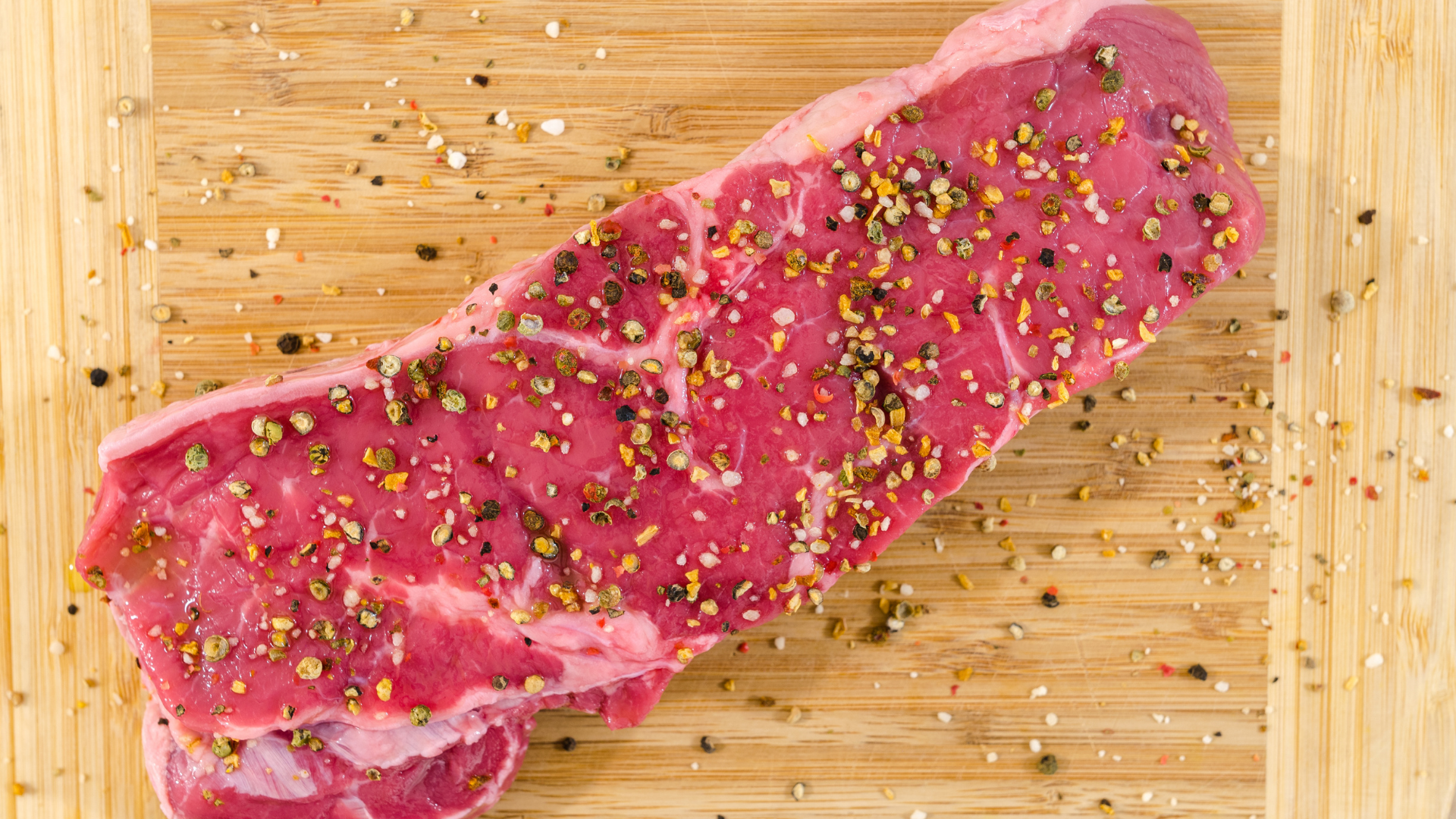 A raw, seasoned beef steak rests on a light-colored wooden cutting board scattered with peppercorns.