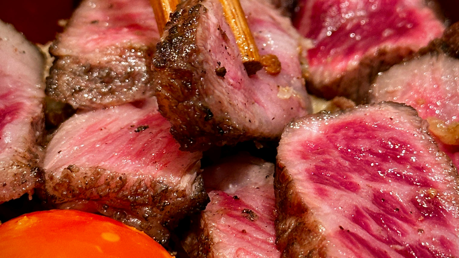Close-up of succulent, medium-rare cubed steak being held by wooden chopsticks next to a bright orange cherry tomato.