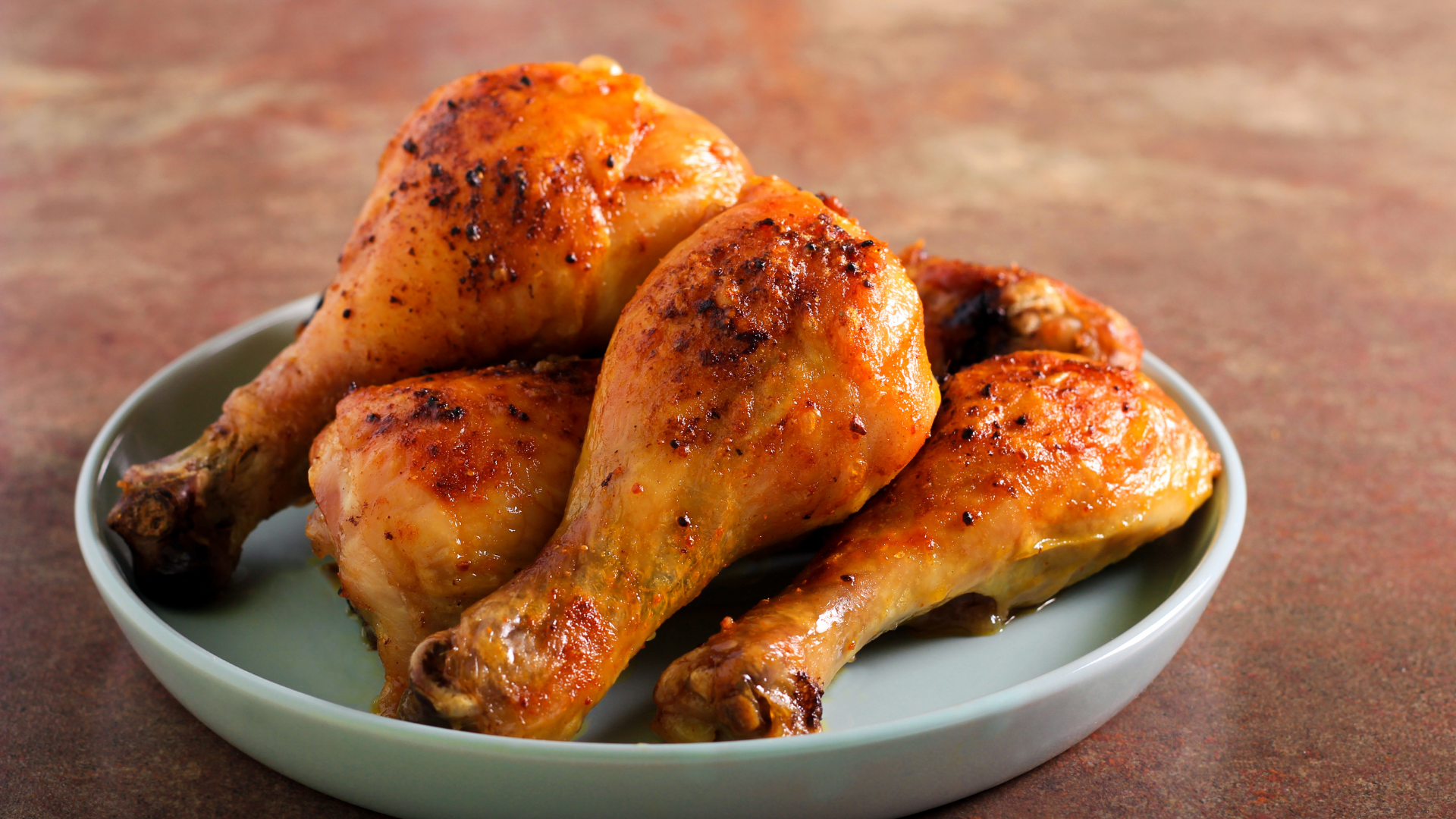 A plate of golden-brown, roasted chicken drumsticks with visible seasoning on a mottled brown surface.