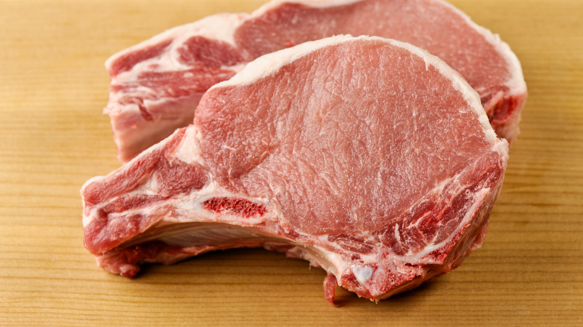 Two raw, bone-in pork chops resting on a light-colored wooden surface.