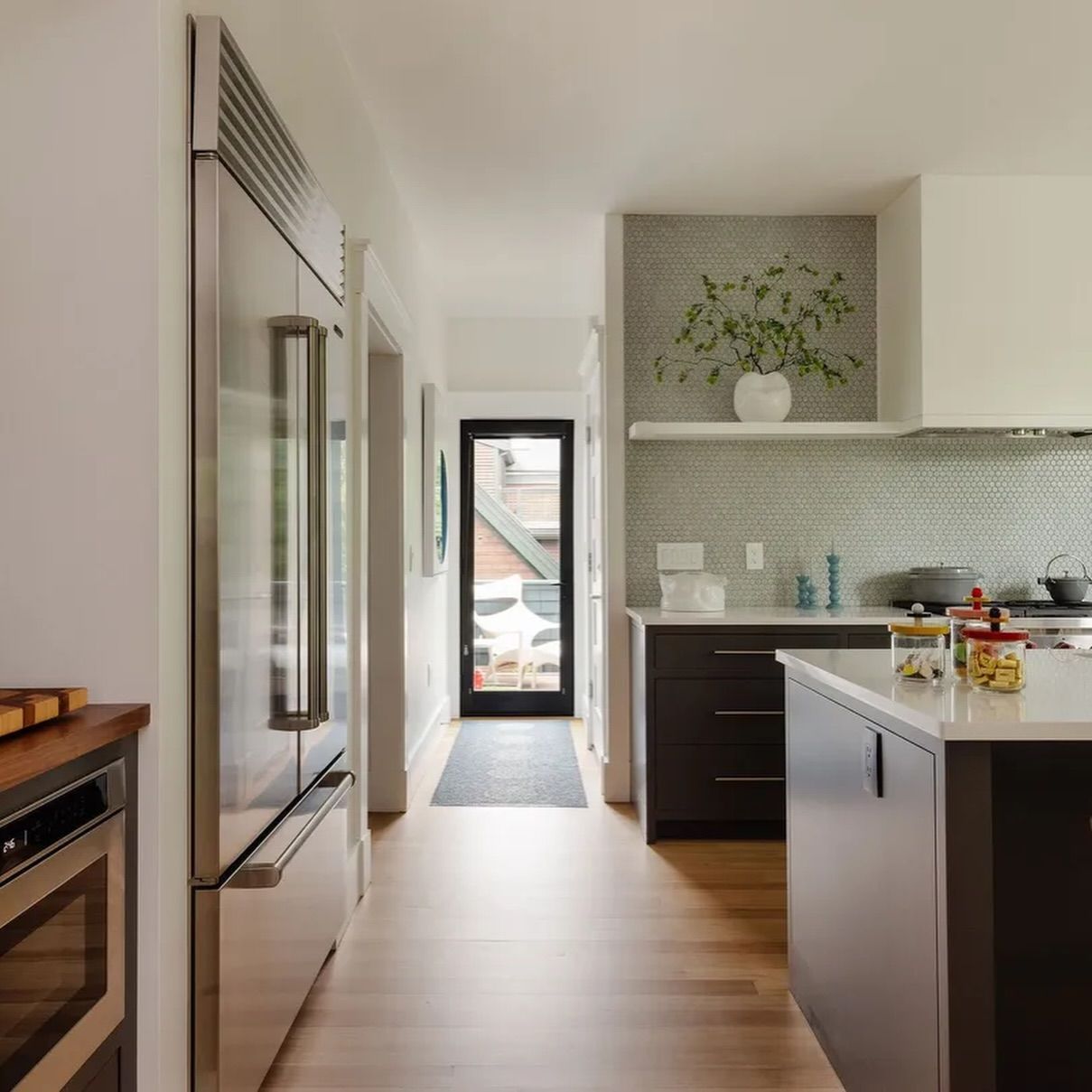 Modern kitchen with stainless steel appliances, dark cabinets, and a bright hallway to a glass door.