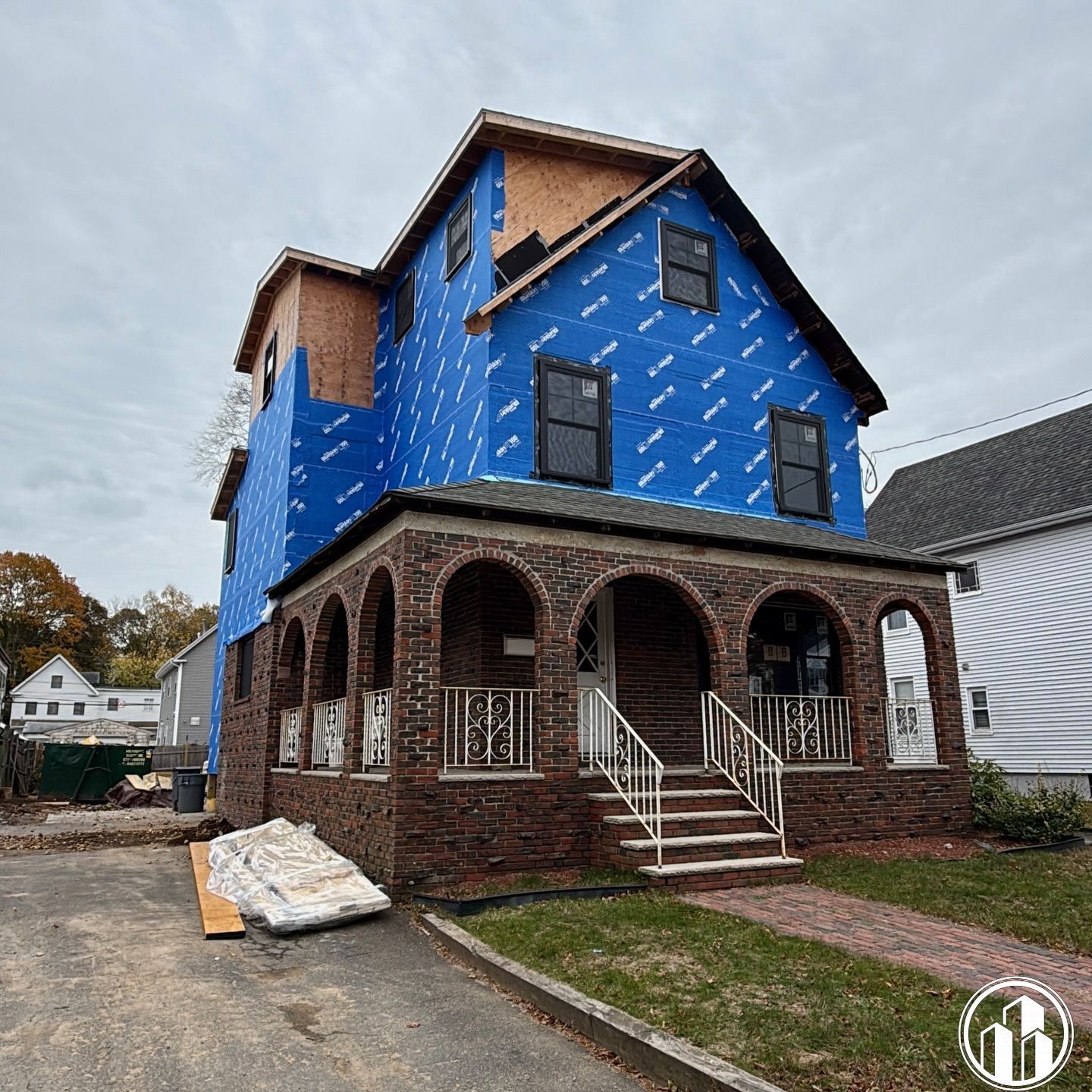 Three-story house under construction with blue house wrap and brick porch, viewed from the driveway