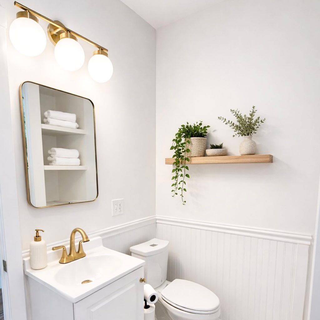 Bright modern bathroom with gold fixtures, white vanity, toilet, mirror, and floating shelf with plants.