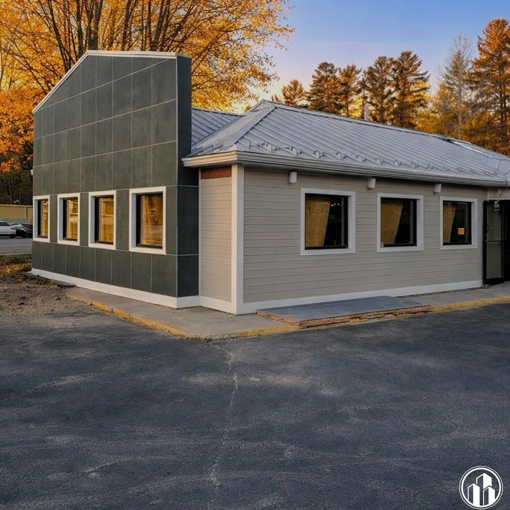 Modern gray building with large windows and a sloped metal roof beside autumn trees at dusk