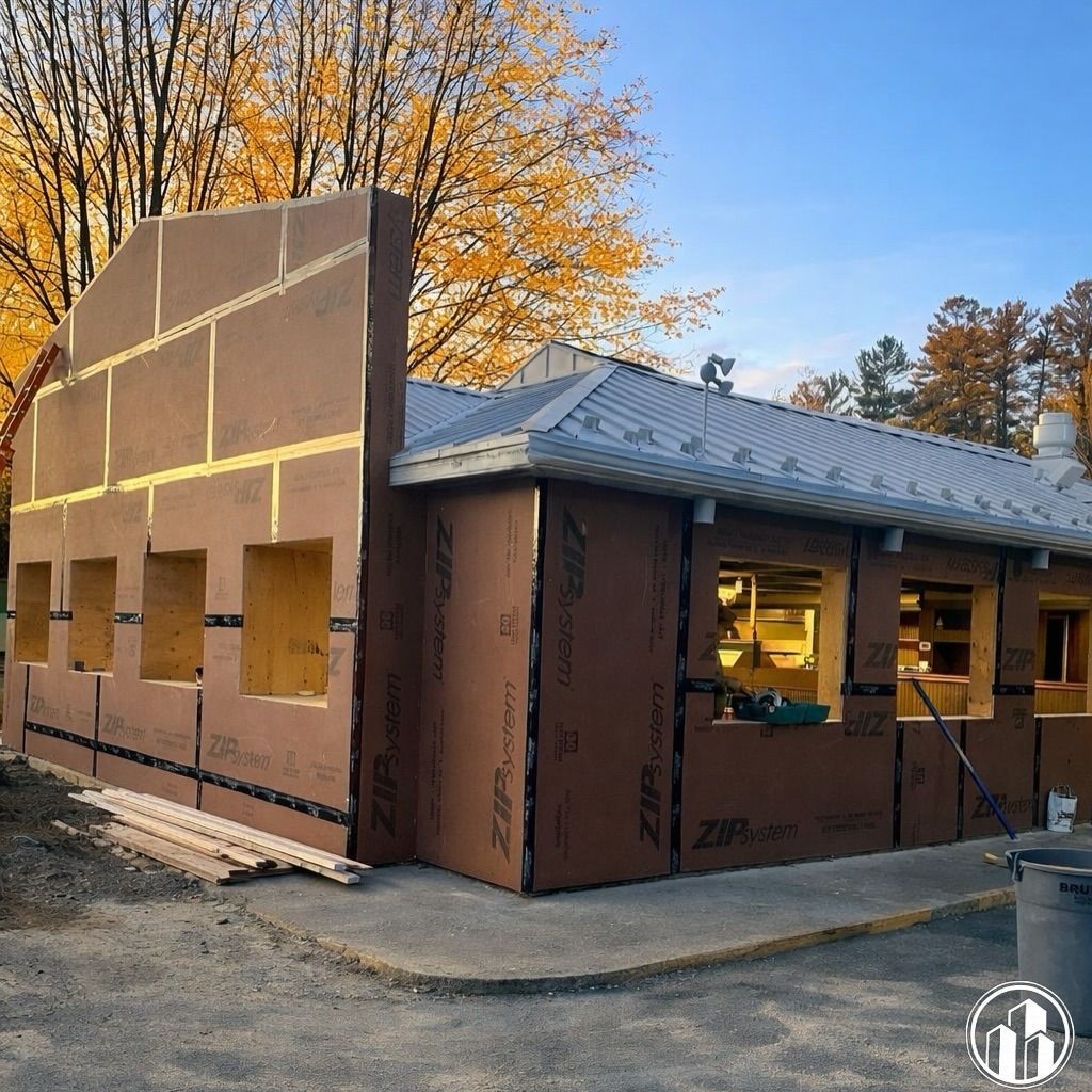 House under construction with brown sheathing, unfinished windows, and autumn trees in the background