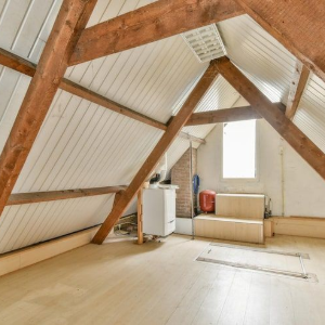 Empty attic room with exposed wooden beams and sloped white walls, lit by a small window.