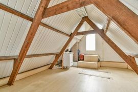 Attic room with exposed wooden beams, white walls, and a small window, mostly empty.