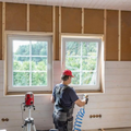 Person painting a room under construction beside two windows and exposed wall studs