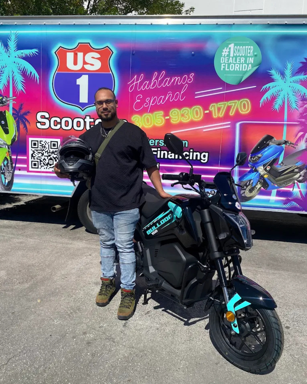 Man holding helmet next to a black scooter in front of a US 1 Scooters truck.