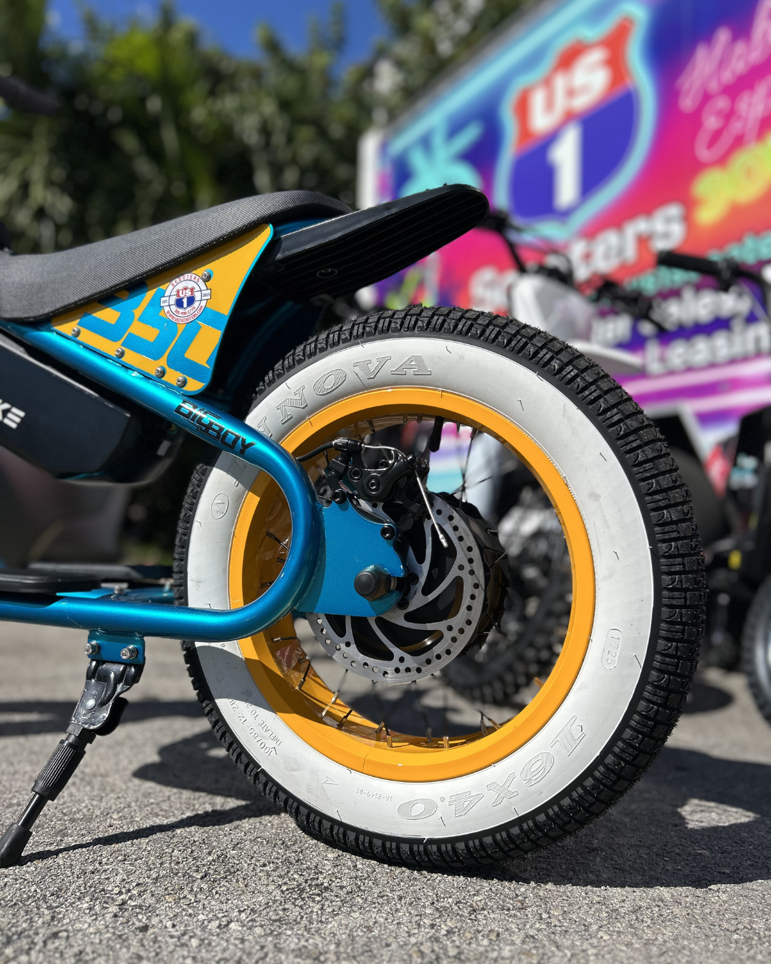 Blue electric bike with yellow and white tire, parked on pavement, US 1 Scooters background.