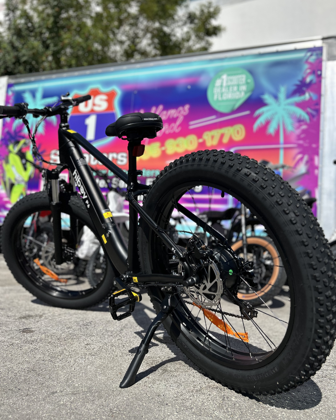 Black fat tire electric bicycle parked on pavement with a colorful trailer in the background.